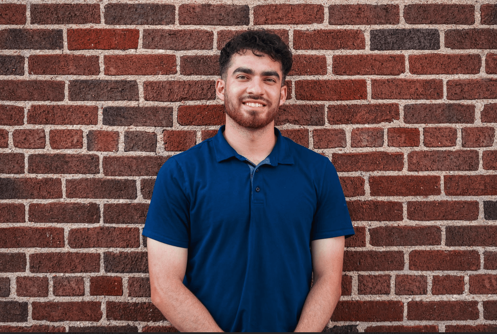 Smiling bearded man in a blue polo shirt standing against a red brick wall.